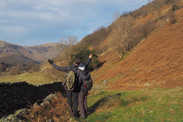 Photograph of a young couple with the backs turned to the camera, arms in the air, standing in the middle of the Lake District surrounded by trees and mountains